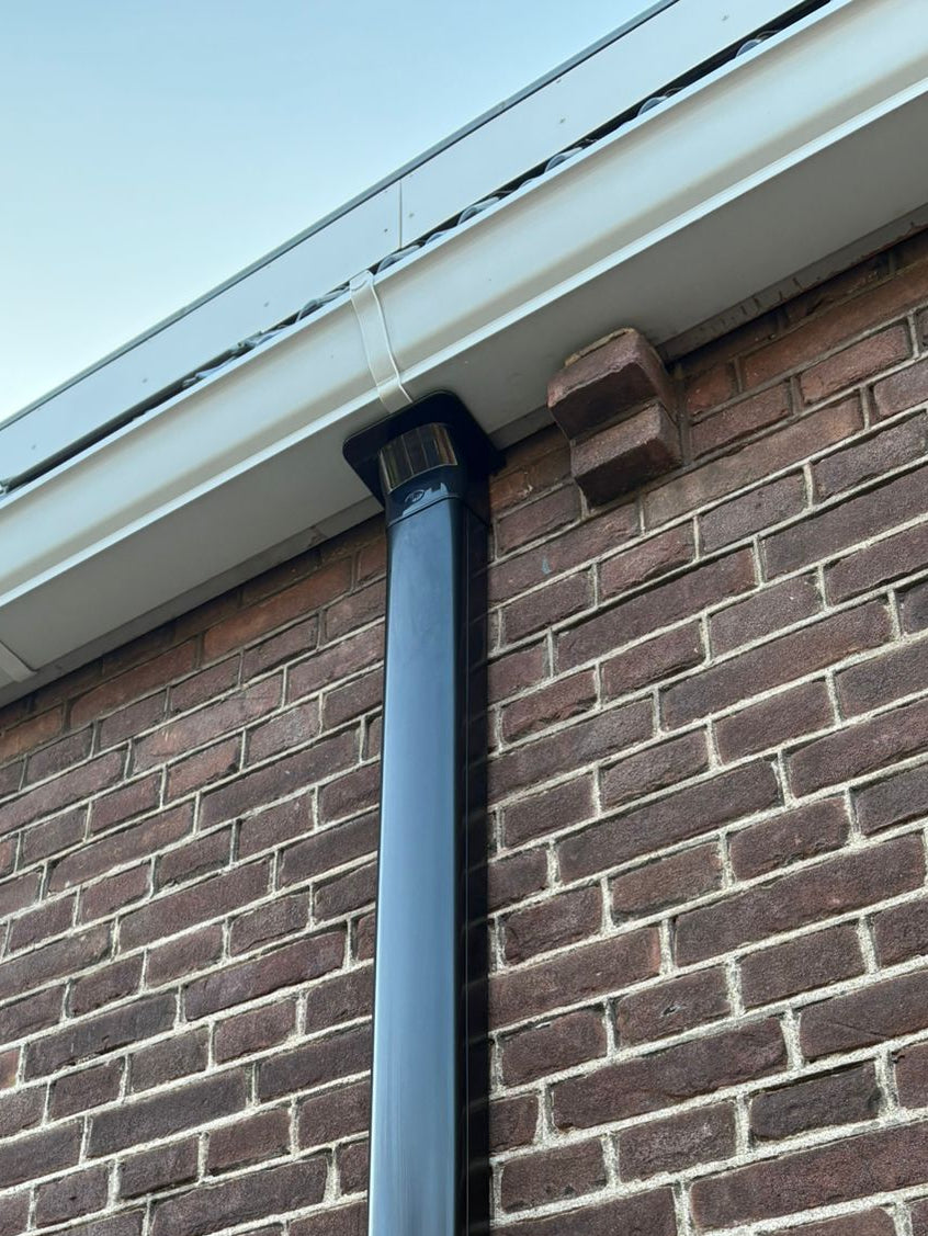 Zwarte Tecnosystemi afwerkgoot (RAL 9005) op bakstenen muur voor airco leidingen, detail bij dakrand/goot.