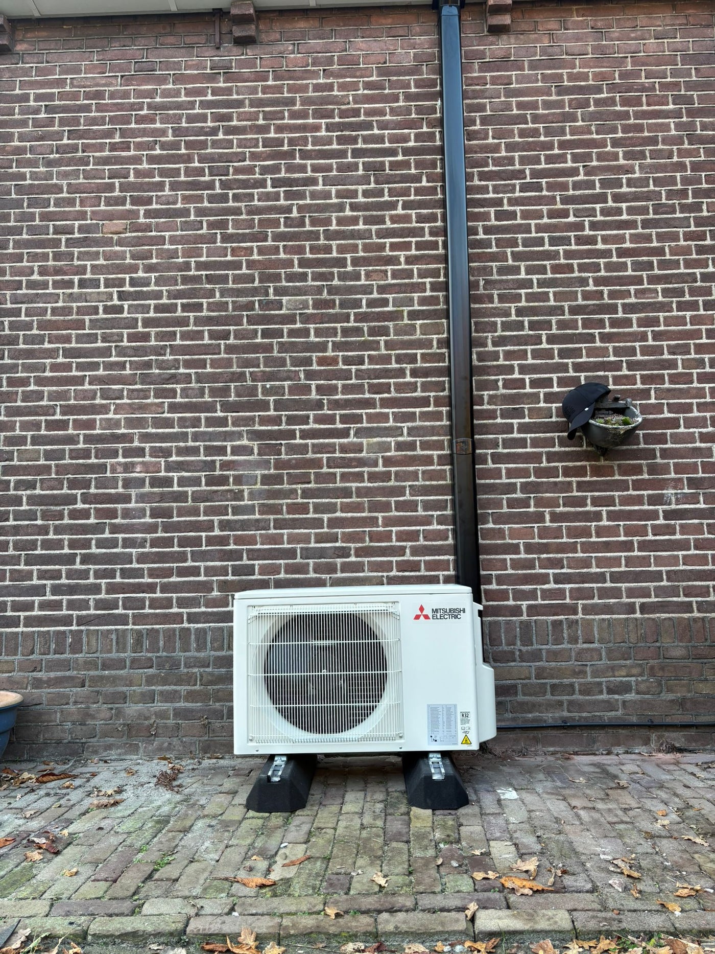 Complete airco-installatie met Mitsubishi Electric buitenunit op rubberen opstelbalken en zwarte Tecnosystemi afwerkgoot (RAL 9005) op bakstenen gevel.