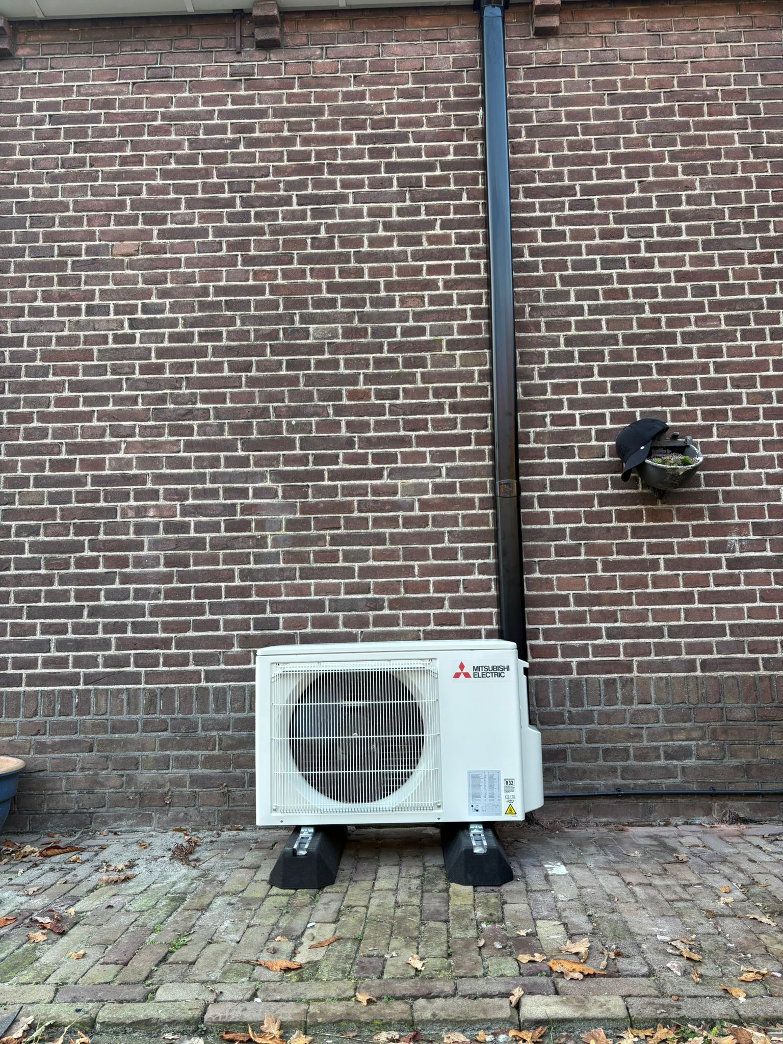 Complete airco-installatie met Mitsubishi Electric buitenunit op rubberen opstelbalken en zwarte Tecnosystemi afwerkgoot (RAL 9005) op bakstenen gevel.
