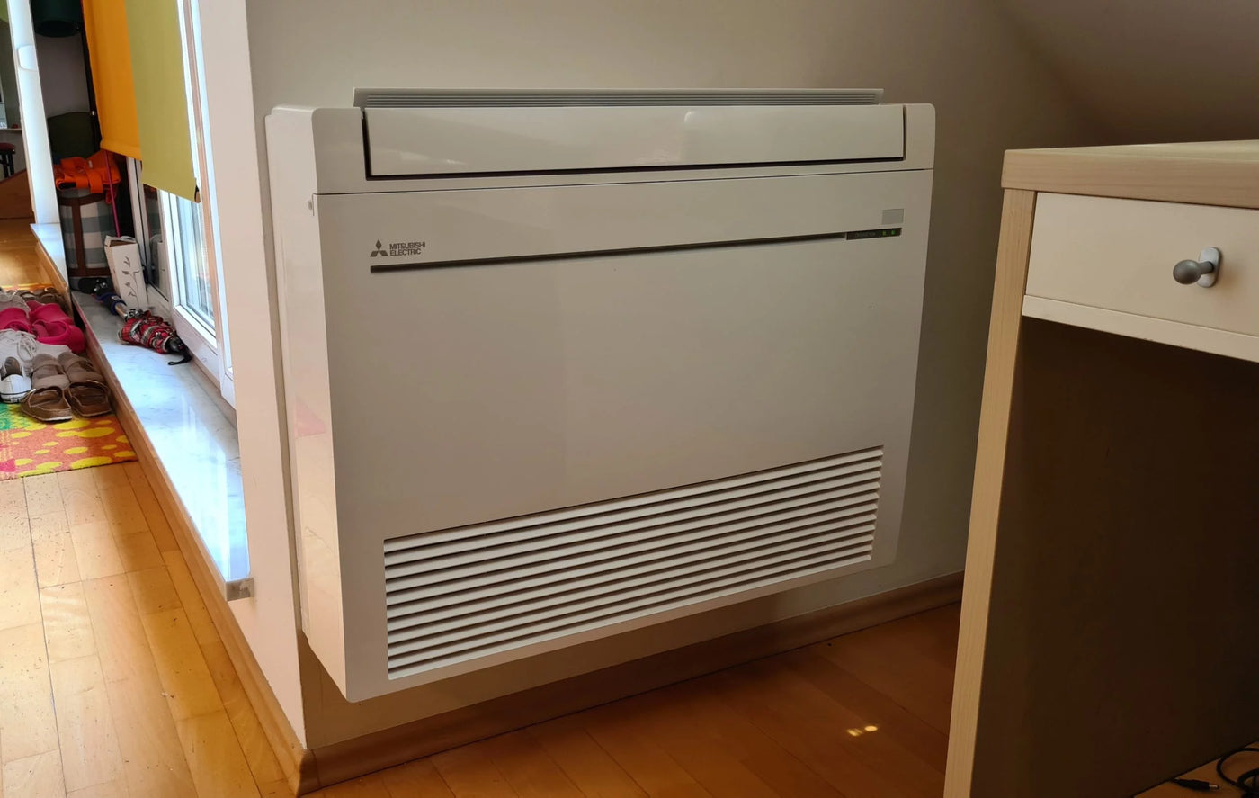 Geïnstalleerde Mitsubishi Electric Vloermodel Binnenunit (Airco/Warmtepomp) gemonteerd tegen een muur in een woon- of slaapkamer. Toont de compacte vloeropstelling voor minimale ruimte-inname.